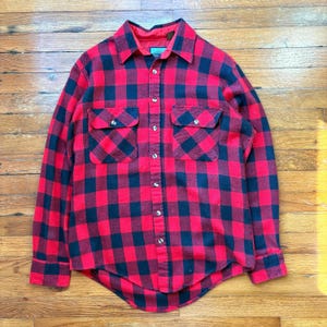 May include: Red and black plaid flannel shirt with two chest pockets and button-down closure. The shirt has a classic lumberjack style, with a collared neckline and long sleeves. The fabric appears to be a soft, warm material.