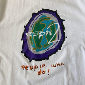 May include: A white t-shirt featuring a graphic of a stylised green and blue earth globe surrounded by a grey and purple border. The text "esprit" is written in white across the globe, and the orange text "people who do!" is printed below.