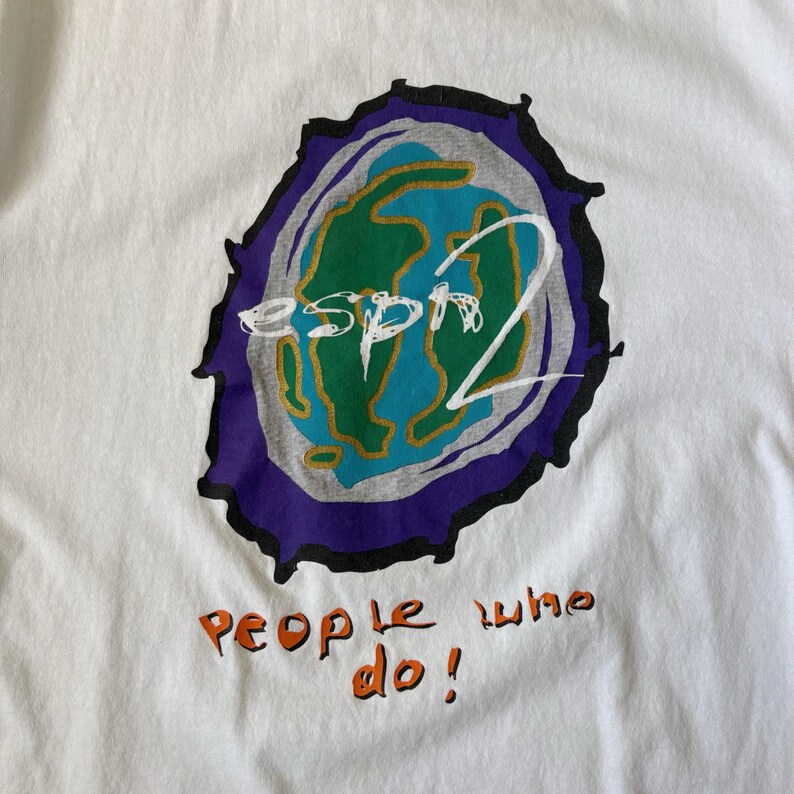 May include: A white t-shirt featuring a graphic of a stylised green and blue earth globe surrounded by a grey and purple border. The text "esprit" is written in white across the globe, and the orange text "people who do!" is printed below.