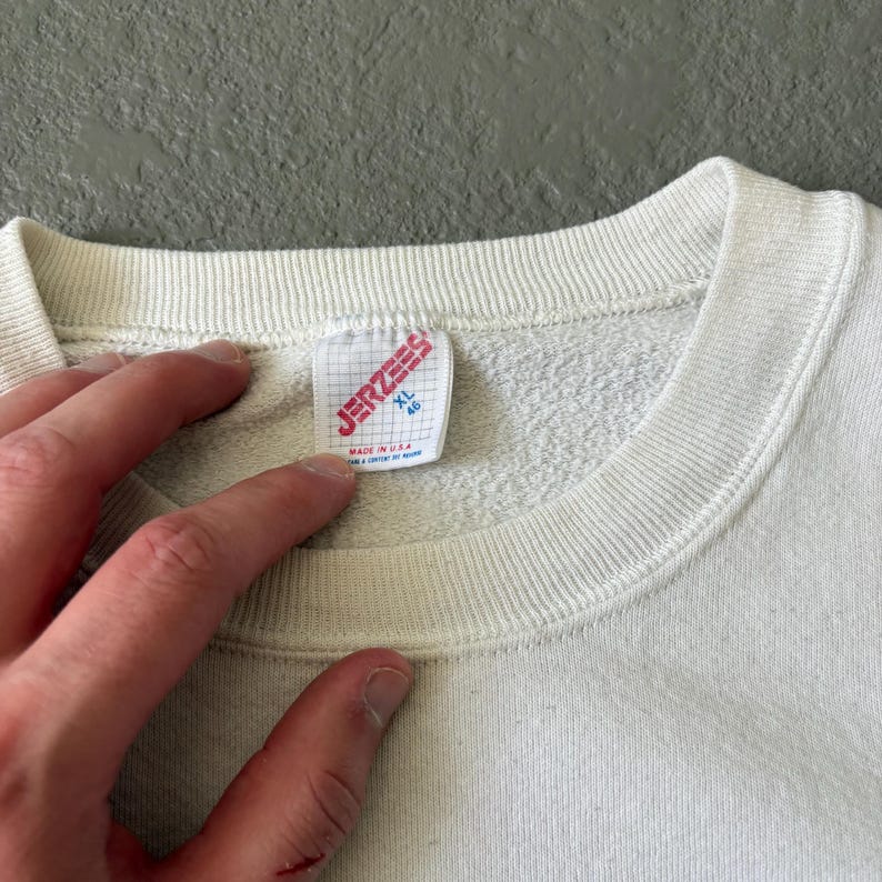 May include: A white crewneck sweatshirt featuring a ribbed collar and a white fabric tag sewn into the inner neckline. The tag displays the red text JERZEES, the size XL 46, and the text MADE IN U.S.A. in red, indicating the garment's origin.