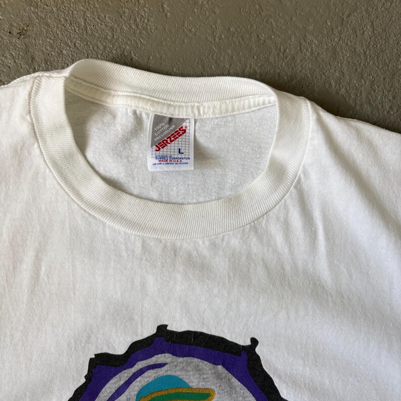 May include: A white short-sleeved t-shirt with a ribbed crew neckline. A white fabric label is sewn inside the collar, displaying the red text JERZEES, the size L, and the phrase 100% COTTON PRESHRUNK MADE IN U.S.A. A partial graphic is visible below.
