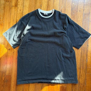 May include: A black short-sleeved t-shirt with a white collar and sleeve trim. The shirt is made of a soft, comfortable material and is suitable for casual wear. The t-shirt is laid flat on a wooden surface.