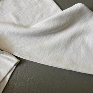 May include: A light gray sweatshirt featuring a soft, fleeced texture and long sleeves with ribbed cuffs. The fabric displays subtle, faint yellowish discoloration spots across the surface, visible on the folded sleeve and main body section.