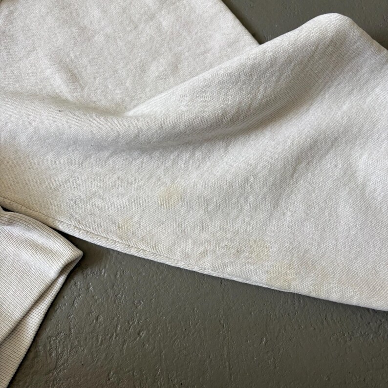 May include: A light gray sweatshirt featuring a soft, fleeced texture and long sleeves with ribbed cuffs. The fabric displays subtle, faint yellowish discoloration spots across the surface, visible on the folded sleeve and main body section.