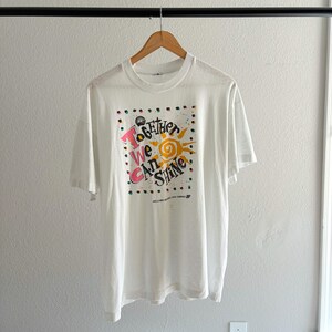 May include: White t-shirt with a colorful graphic that reads "Together We Can Shine." The design includes a sun, dots, and the text in pink, yellow, and blue. The shirt is hanging on a wooden hanger.