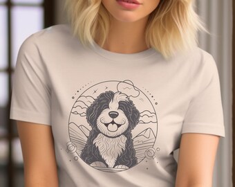 May include: Beige t-shirt featuring a graphic of a smiling black and white dog within a circular design. The design includes mountains, clouds, and decorative elements. The shirt is made of a soft material.
