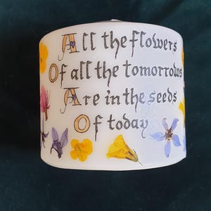 May include: A white cylindrical candle decorated with pressed yellow, pink, and blue flowers. The text printed in black ink reads: All the flowers Of all the tomorrows Are in the seeds Of today. The candle has a smooth surface and a flat top.