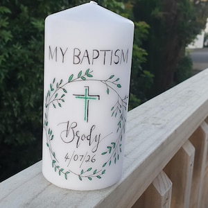 May include: A white pillar candle featuring the text MY BAPTISM at the top, a central green cross inside a hand-drawn green leaf wreath, and the name Brody with the date 4/07/26 written in black script lettering on the front surface.