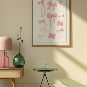May include: A framed print of a watercolour illustration of pink bows in various styles and sizes. The print is hanging on a light beige wall with a green vase and a pink lamp on a wooden table in the foreground.