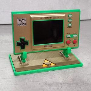 Könnte beinhalten: Eine grüne und goldene Game & Watch Handheld-Videospielkonsole mit The Legend of Zelda. Die Konsole steht auf einem grünen Ständer mit einem goldenen Triforce-Symbol.