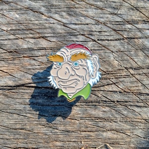 May include: A colorful enamel pin featuring a grumpy-looking troll face. The pin has a red cap, yellow eyebrows, blue eyes, and a green collar. The pin is set against a weathered wood background.