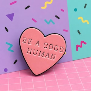 May include: A pink heart-shaped enamel pin with the words "BE A GOOD HUMAN" in black lettering. The pin has a black border and is set against a background with geometric shapes in pink, purple, and teal.