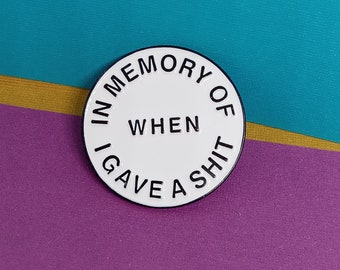 May include: A round, white enamel pin with black text. The text reads "IN MEMORY OF WHEN I GAVE A SHIT" in a circular pattern around the edge, with "WHEN" in the center. The pin has a black border and is set against a teal, gold, and purple background.