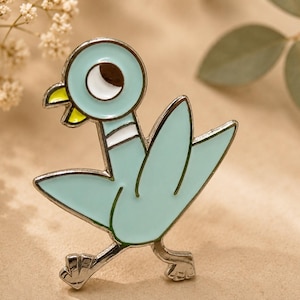 May include: A light blue enamel pin featuring a cartoon pigeon with a silver outline. The pigeon has a round head with a brown and white eye, yellow beak, and white collar. The pin is a whimsical accessory.