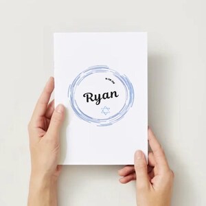 May include: A white card featuring the name "Ryan" in black script, surrounded by a blue watercolour design. A Star of David is beneath the name. The card is held by two hands against a white backdrop.