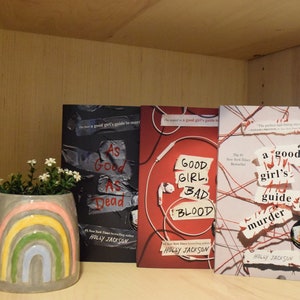 May include: Three paperback books with the title "A Good Girl's Guide to Murder" by Holly Jackson. The books are stacked on a wooden shelf. The book covers are red, white, and black with text in white and red.