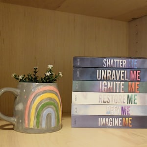 May include: A stack of six paperback books with the titles "Shatter Me", "Unravel Me", "Ignite Me", "Restore Me", "Defy Me", and "Imagine Me". The books are all by the same author, Tahereh Mafi. A gray ceramic mug with a rainbow design is in the foreground.