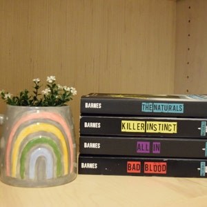 May include: A stack of five black paperback books with white text on the spines. The titles are "The Naturals", "Killer Instinct", "All In", and "Bad Blood". The books are on a wooden shelf with a gray ceramic mug with a rainbow design and white flowers in front of them.