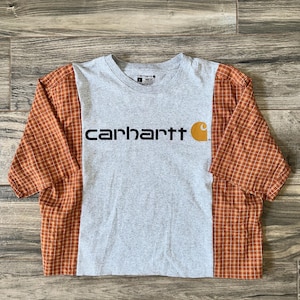 Könnte beinhalten: Graues Carhartt-T-Shirt mit orangefarbenen karierten Ärmeln. Das Carhartt-Logo ist schwarz mit einem orangefarbenen 'C' auf der rechten Seite gedruckt. Das Shirt ist ein Cropped-Style.
