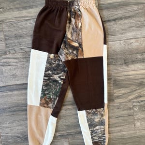 May include: Patchwork fleece sweatpants in brown, tan, white, and camouflage patterns. The pants have a drawstring waist and elastic cuffs. The design features a mix of solid colors and a woodland camouflage print.