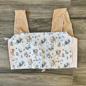 May include: A handmade, light pink and white top with a Bambi-themed print. The top features a square neckline and wide straps in a soft, light brown color. The fabric is patterned with deer, squirrels, and floral elements.