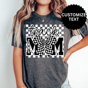 May include: Dark gray t-shirt with a checkered design and the words "track MOM" in a stylized font. A large checkered bow is centered on the design. The name "EMMA" is vertically aligned on the left side of the "M".