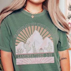 May include: Green t-shirt with a vintage-style graphic featuring a mountain range, cacti, and the text "His Mercies Are New Every Morning" and "Lamentations 3:23".