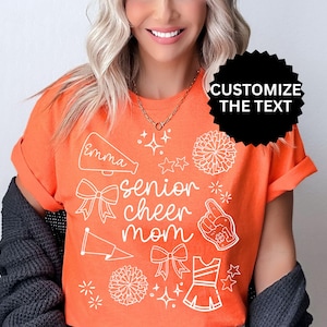 May include: Orange t-shirt with white graphics, including a megaphone, bows, stars, pom-poms, and a cheerleader uniform. The text "Emma" and "senior cheer mom" are also visible. The shirt has rolled-up sleeves.