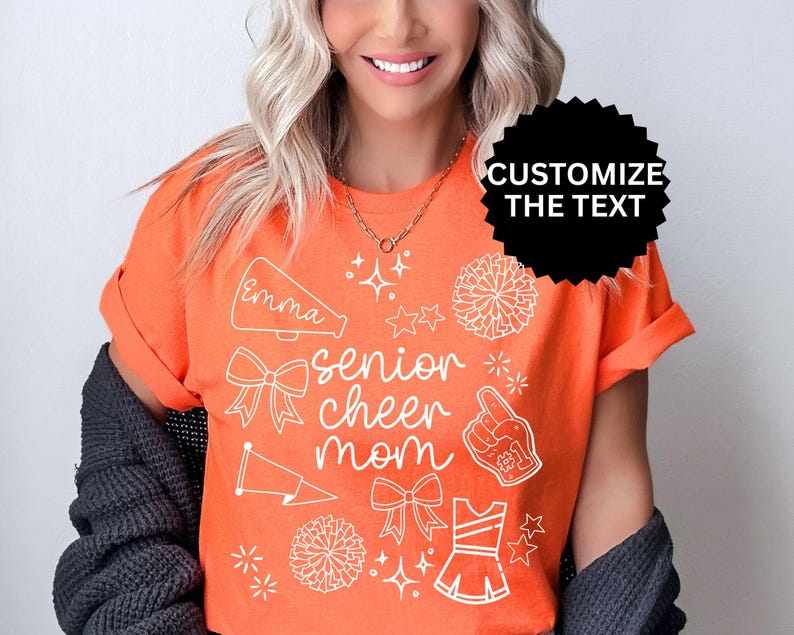 May include: Orange t-shirt with white graphics, including a megaphone, bows, stars, pom-poms, and a cheerleader uniform. The text "Emma" and "senior cheer mom" are also visible. The shirt has rolled-up sleeves.