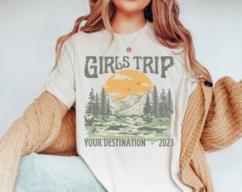 Op de afbeelding: Crèmekleurig T-shirt met een retro grafisch ontwerp. Het ontwerp toont de tekst "GIRLS TRIP" boven een berg- en boslandschap met een ondergaande zon. Daaronder staat de tekst "YOUR DESTINATION - 2023".