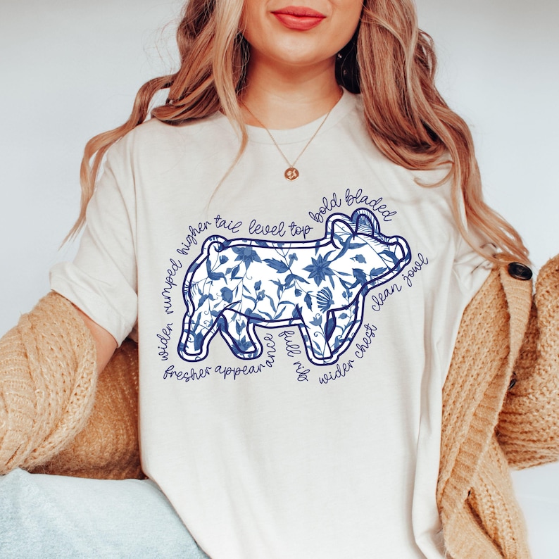 May include: Cream-colored t-shirt featuring a blue floral pig design with text. The pig graphic is surrounded by phrases like "wider rumped higher" and "clean jowl". The shirt is a casual, everyday wear item.