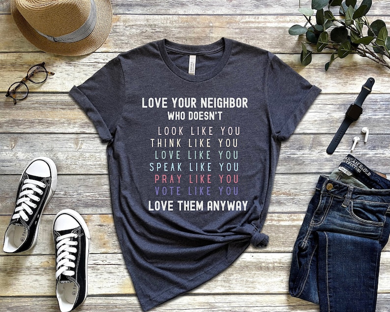 Op de afbeelding: Een donkergrijs T-shirt met de tekst "LOVE YOUR NEIGHBOR WHO DOESN'T LOOK LIKE YOU THINK LIKE YOU LOVE LIKE YOU SPEAK LIKE YOU PRAY LIKE YOU VOTE LIKE YOU LOVE THEM ANYWAY" gedrukt in kleurrijke letters.