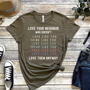 Op de afbeelding: Een groen T-shirt met de tekst "Lief je buurman die niet... eruitziet als jij, denkt als jij, van je houdt, praat als jij, bidt als jij, stemt als jij, hou van hem toch".