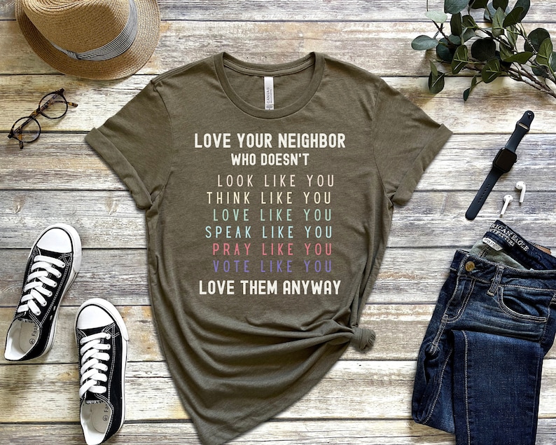 Op de afbeelding: Een groen T-shirt met de tekst "Lief je buurman die niet... eruitziet als jij, denkt als jij, van je houdt, praat als jij, bidt als jij, stemt als jij, hou van hem toch".