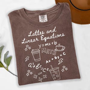 May include: A brown t-shirt with the words "Lattes and Linear Equations" in white script. The shirt features math equations and coffee-related graphics, including a coffee cup and a to-go cup. The shirt is made by Comfort Colors.