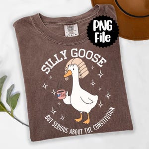 Könnte beinhalten: Braunes T-Shirt mit dem Text "SILLY GOOSE" und einer Cartoon-Gans, die eine Perücke trägt und eine Kaffeetasse mit "USA" darauf hält. Der Text "BUT SERIOUS ABOUT THE CONSTITUTION" steht darunter.