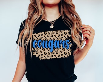 May include: Black t-shirt with a brown and tan leopard print design and the word "cougars" in blue lettering.