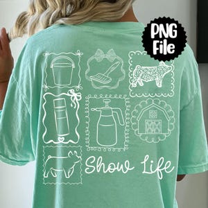May include: A light green t-shirt featuring white line drawings of farm-related items, including a bucket, brush, pig, spray bottle, barn, and the words "Show Life". The shirt has a relaxed fit.
