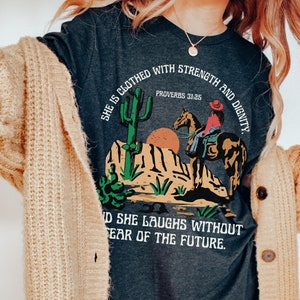 May include: Dark gray t-shirt featuring a desert scene with a cowgirl on horseback, a cactus, and the text "She is clothed with strength and dignity. Proverbs 31:25" and "And she laughs without fear of the future."