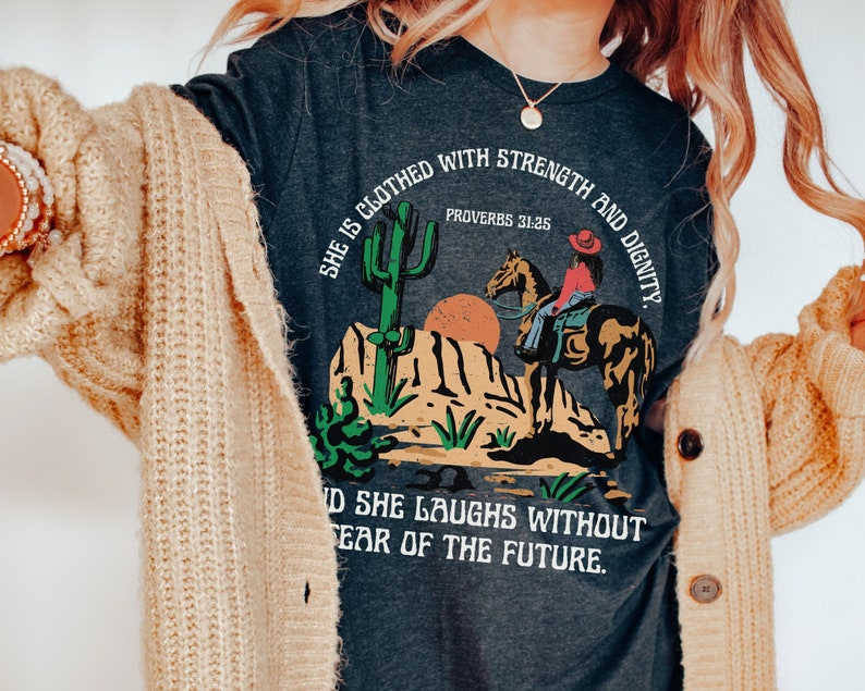 May include: Dark gray t-shirt featuring a desert scene with a cowgirl on horseback, a cactus, and the text "She is clothed with strength and dignity. Proverbs 31:25" and "And she laughs without fear of the future."