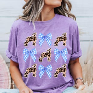 May include: A lilac-colored t-shirt featuring a repeating pattern of leopard-print sheep and blue gingham bows. The sheep are in a standing pose, and the bows are tied in the center of the shirt. The shirt has short sleeves and a relaxed fit.