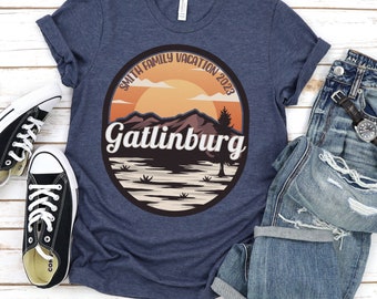 May include: A navy blue t-shirt with a circular graphic featuring a mountain landscape, sunset, and the text "Gatlinburg" and "Smith Family Vacation 2023". The shirt is paired with jeans and black sneakers.