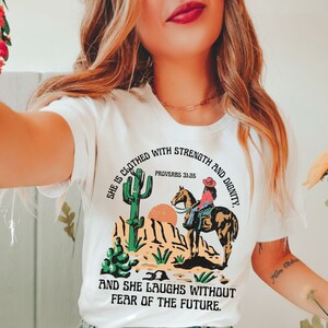 May include: White t-shirt with a desert-themed graphic featuring a cowgirl on horseback, cacti, and mountains. The text reads "She is clothed with strength and dignity" and "And she laughs without fear of the future."