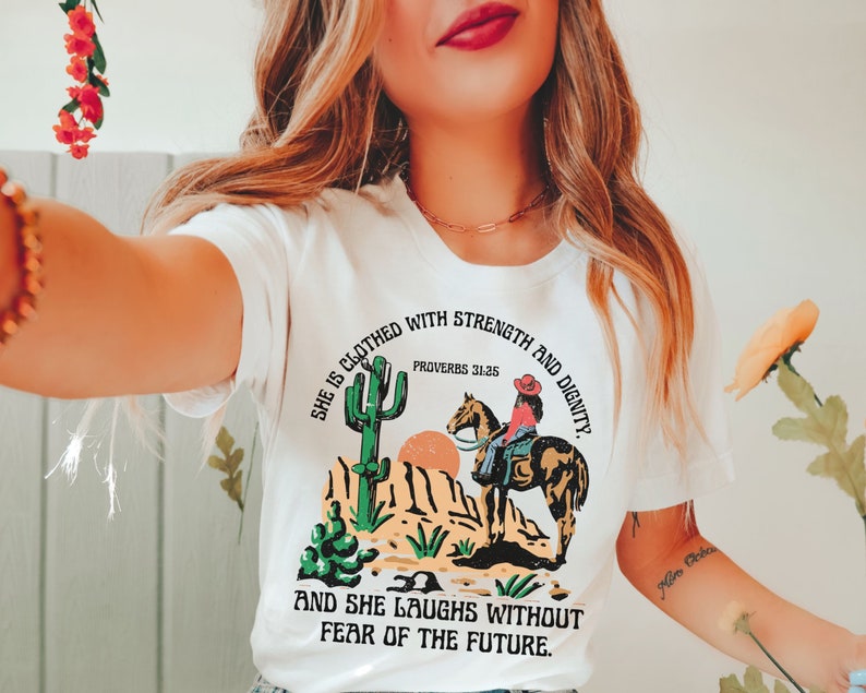 May include: White t-shirt with a desert-themed graphic featuring a cowgirl on horseback, cacti, and mountains. The text reads "She is clothed with strength and dignity" and "And she laughs without fear of the future."