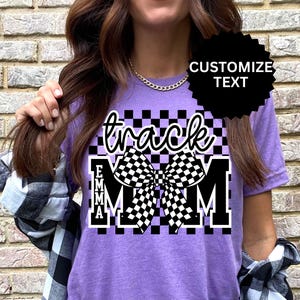 May include: A purple t-shirt with a checkered design and the words "track MOM" in a script font. The name "EMMA" is printed below the word "track". A black and white checkered bow is in the center of the design.