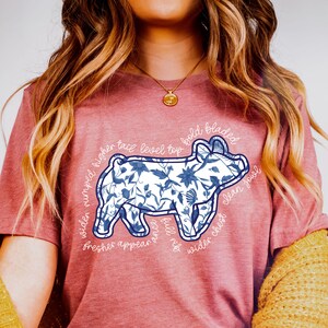 May include: A dusty rose t-shirt featuring a blue and white floral pig design. The pig illustration has text around it, including "wider rumped", "clean jowl", and "fresher appear".