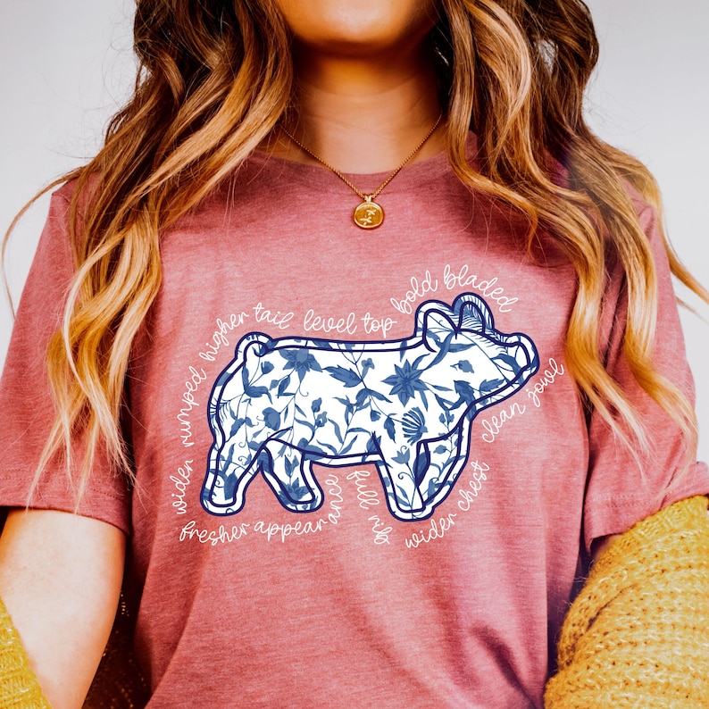 May include: A dusty rose t-shirt featuring a blue and white floral pig design. The pig illustration has text around it, including "wider rumped", "clean jowl", and "fresher appear".