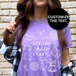May include: A purple t-shirt with white graphics, including a megaphone, bows, stars, a pom-pom, a foam finger, and a cheerleader uniform. The text reads "imma senior cheer mom". The shirt is a casual style.