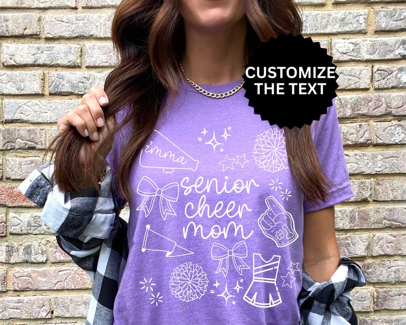 May include: A purple t-shirt with white graphics, including a megaphone, bows, stars, a pom-pom, a foam finger, and a cheerleader uniform. The text reads "imma senior cheer mom". The shirt is a casual style.