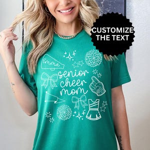 May include: A teal green t-shirt with white graphics, including a megaphone with the name "Emma", bows, stars, pom-poms, a foam finger, and a cheerleader uniform. The text "senior cheer mom" is also printed on the shirt.